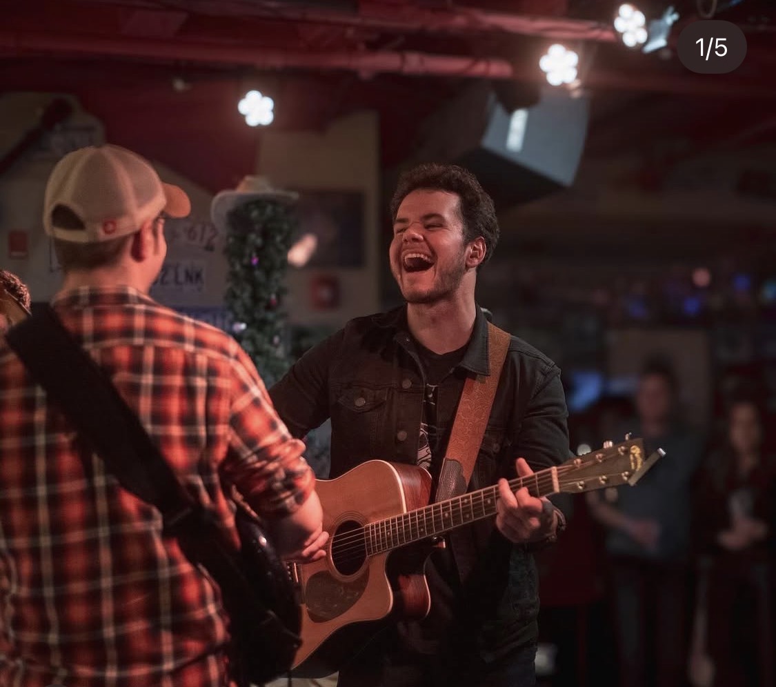 Two people playing guitars and singing in a dimly lit venue, smiling and enjoying music, with a decorated background.