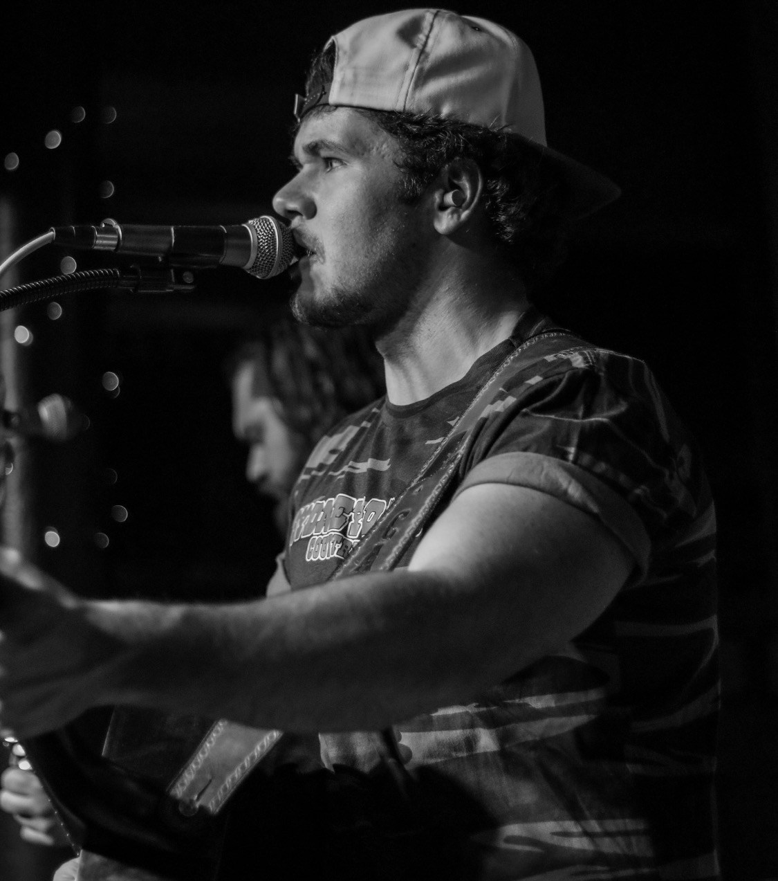 Man singing into microphone, wearing a cap backwards, playing guitar, in a dark setting, black and white photo.