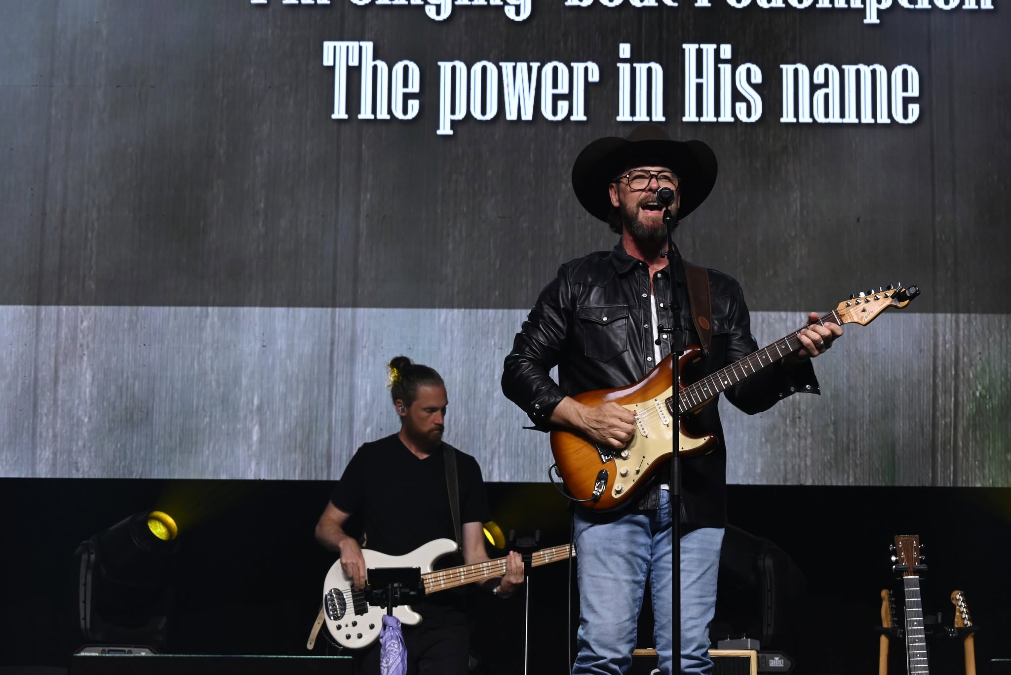A man with a hat and glasses plays an electric guitar on stage, with a woman playing bass behind him.
