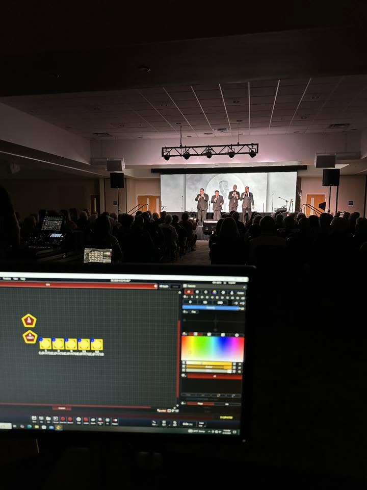 People on stage performing in front of an audience in a large auditorium, with a computer screen in the foreground.