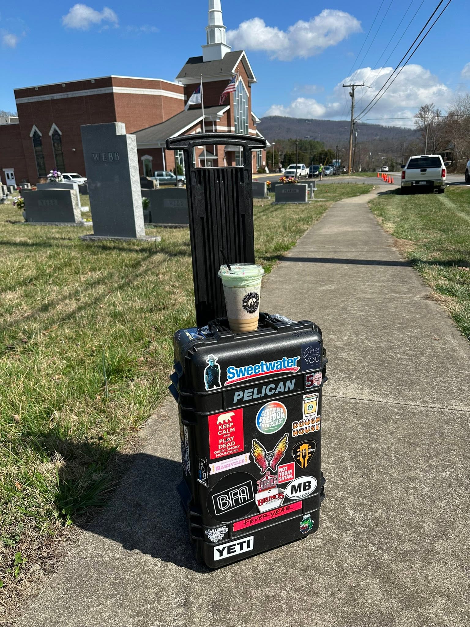 Trash bin with stickers and a coffee cup on top, on a sidewalk near a church and gravestones.