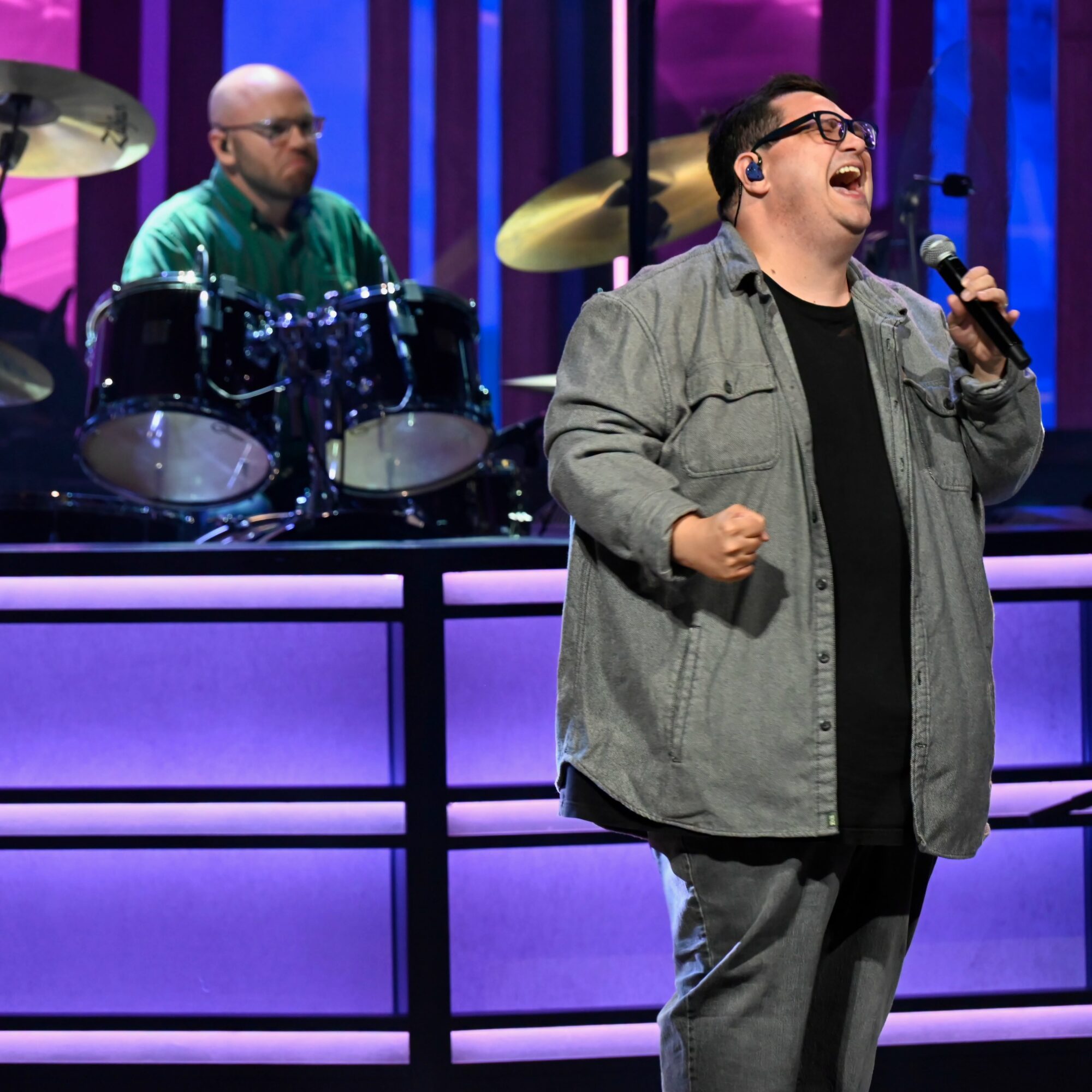 A man singing into a microphone on stage with a drummer in the background, colorful stage lighting, and purple panels.