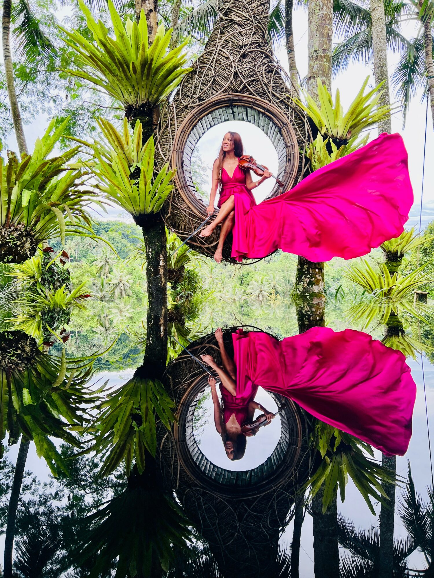 Woman in a pink dress standing in front of a circular wooden structure surrounded by lush greenery, with her reflection in water.