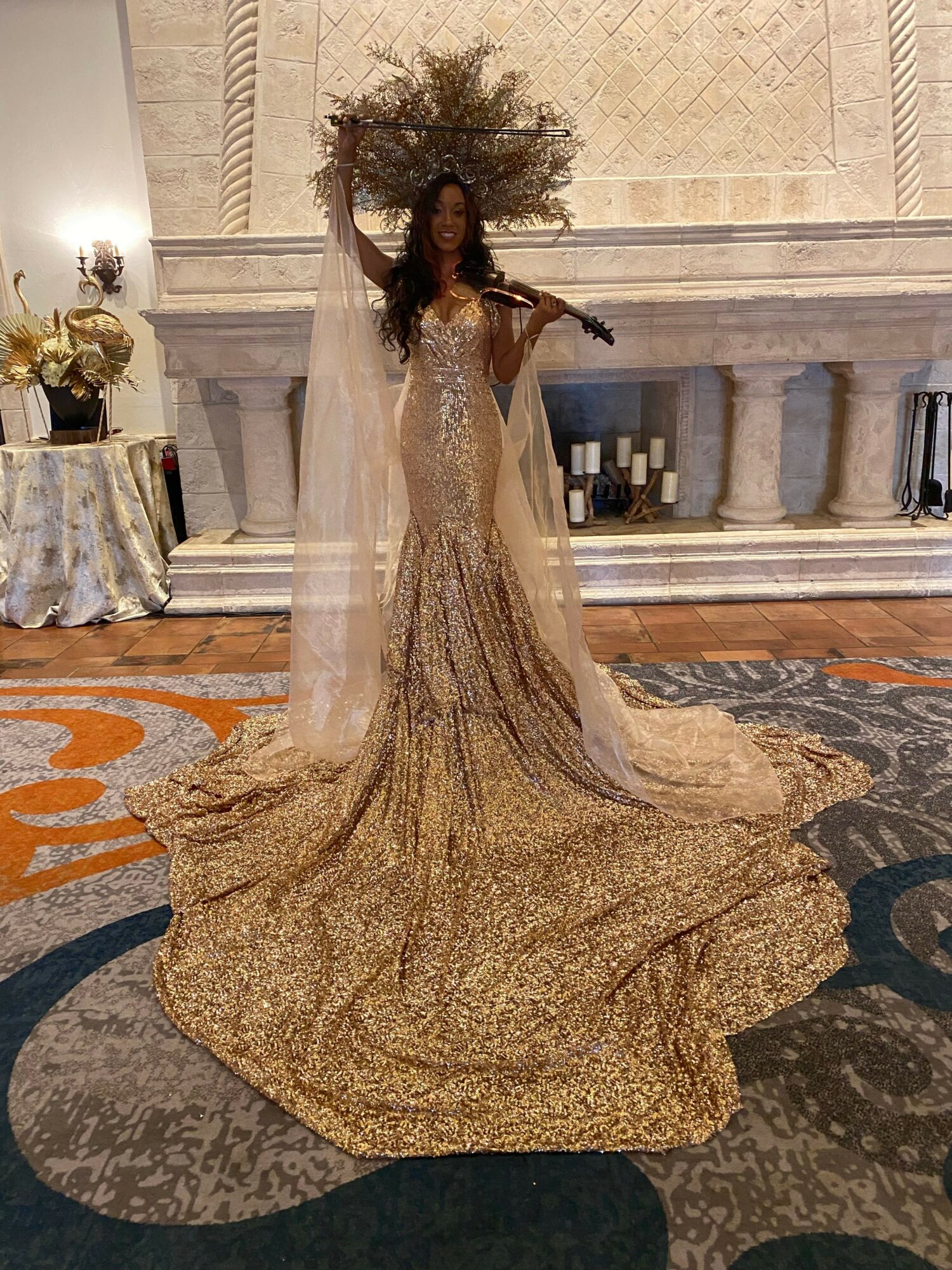 Person in a gold, strapless gown with a long train, standing in a room with patterned carpet and a table with flowers.