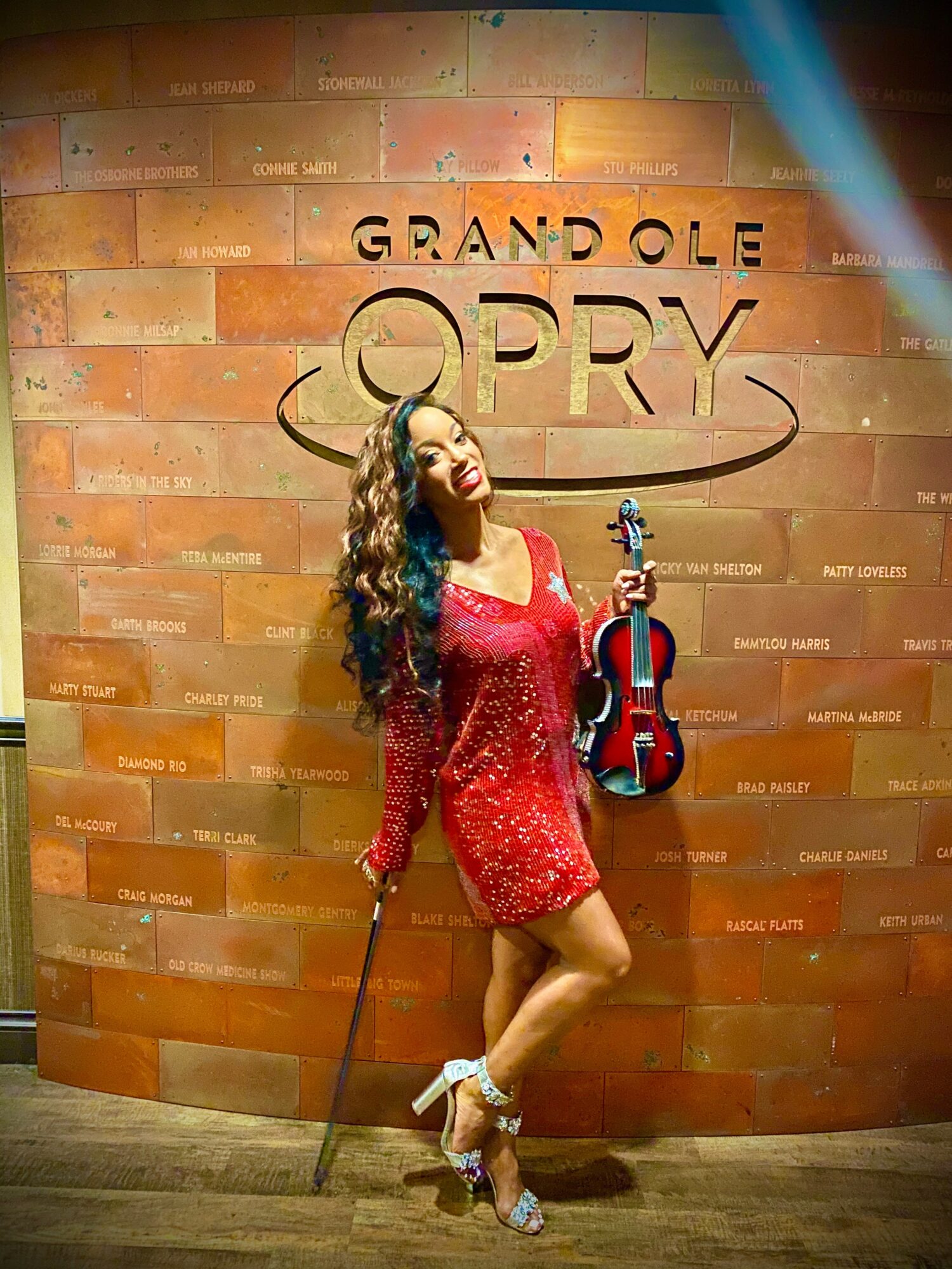 Woman in red dress holding a violin and bow, standing in front of a brick wall with 'Grand Ole Opry' sign.
