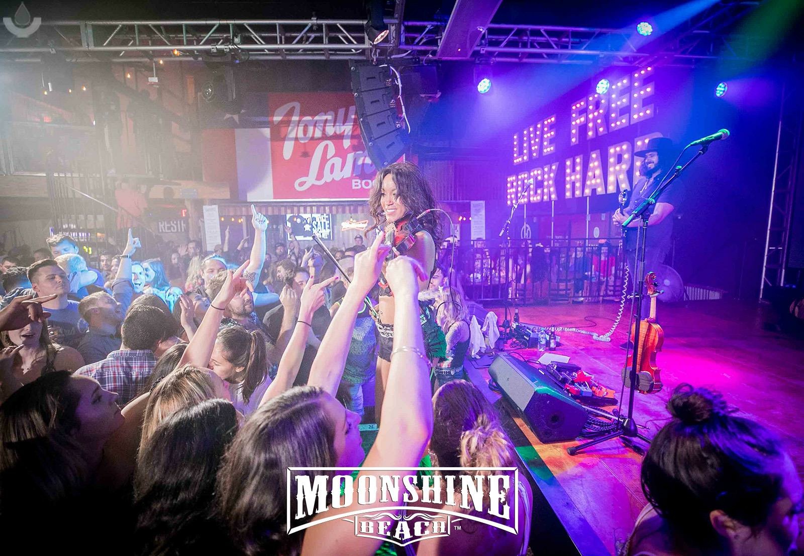 Performer on stage with microphone, audience raising hands, colorful stage lights, and a sign reading 'LOVE PEACE ROCK HARD'.