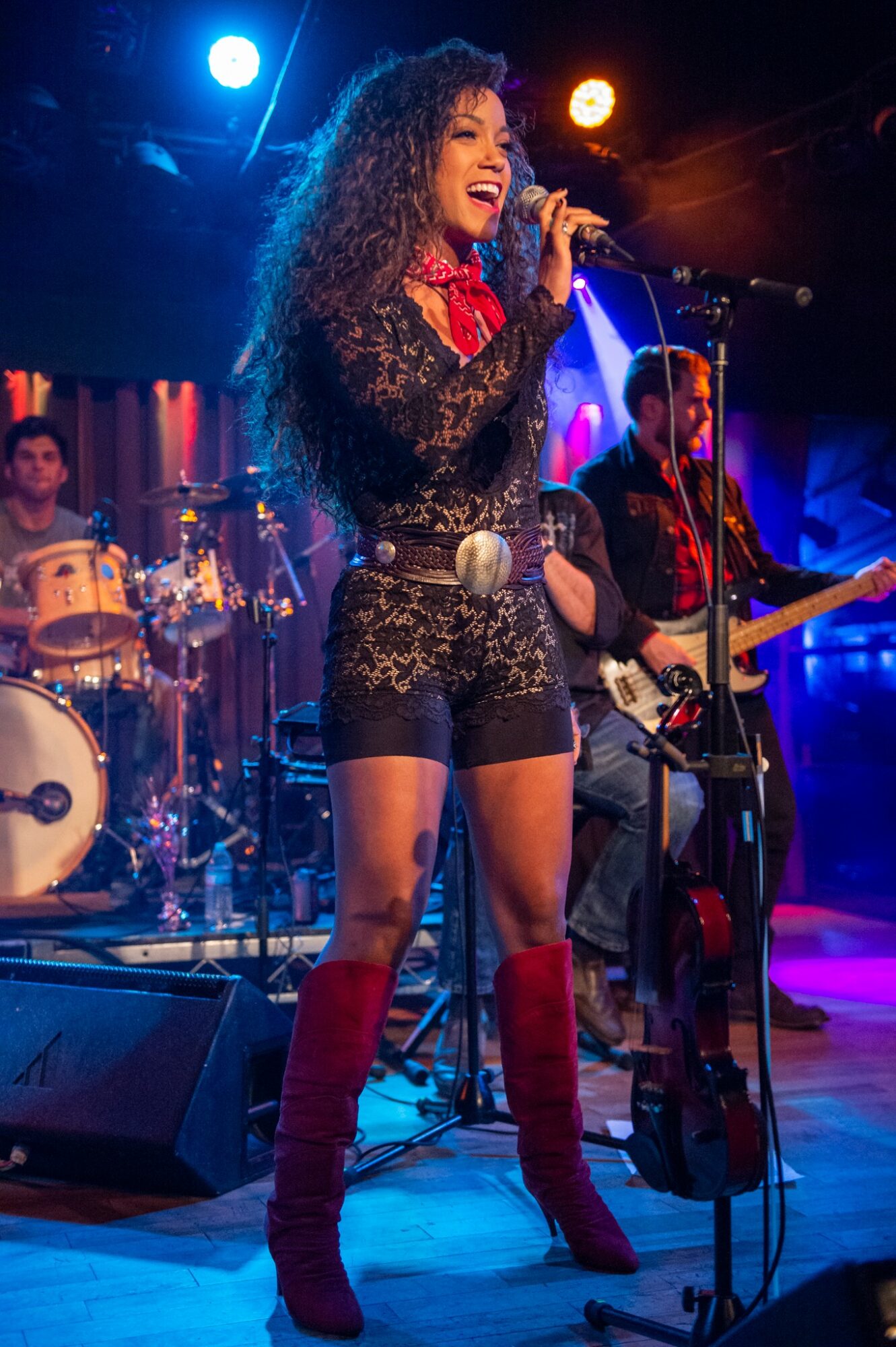 A woman singing into a microphone on stage with band members playing instruments behind her, colorful stage lights shining.