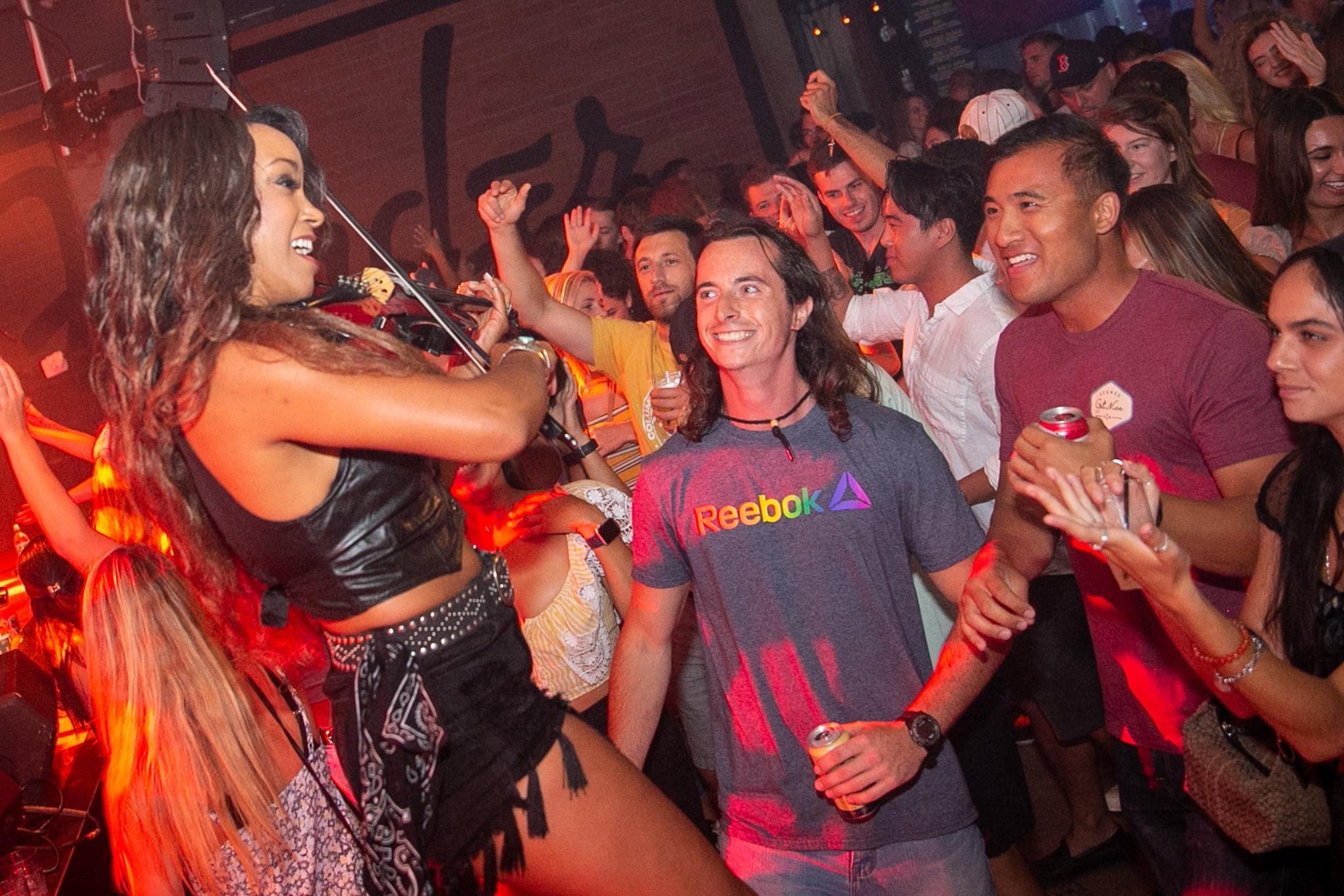 A woman performs on stage with a microphone, audience members watch and smile in a lively indoor setting.