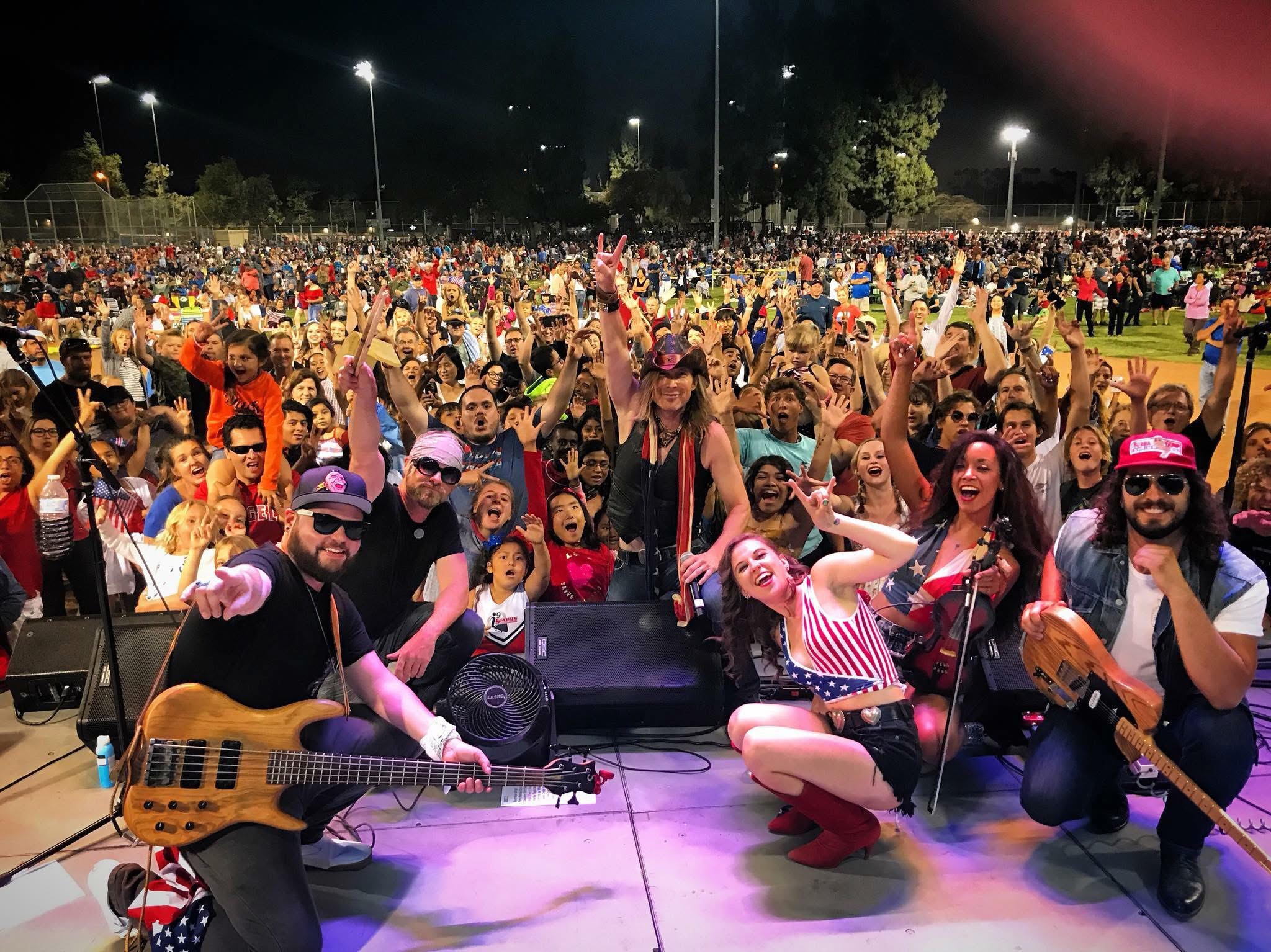 Group of performers on stage with a large crowd at night, some holding instruments and raising hands.
