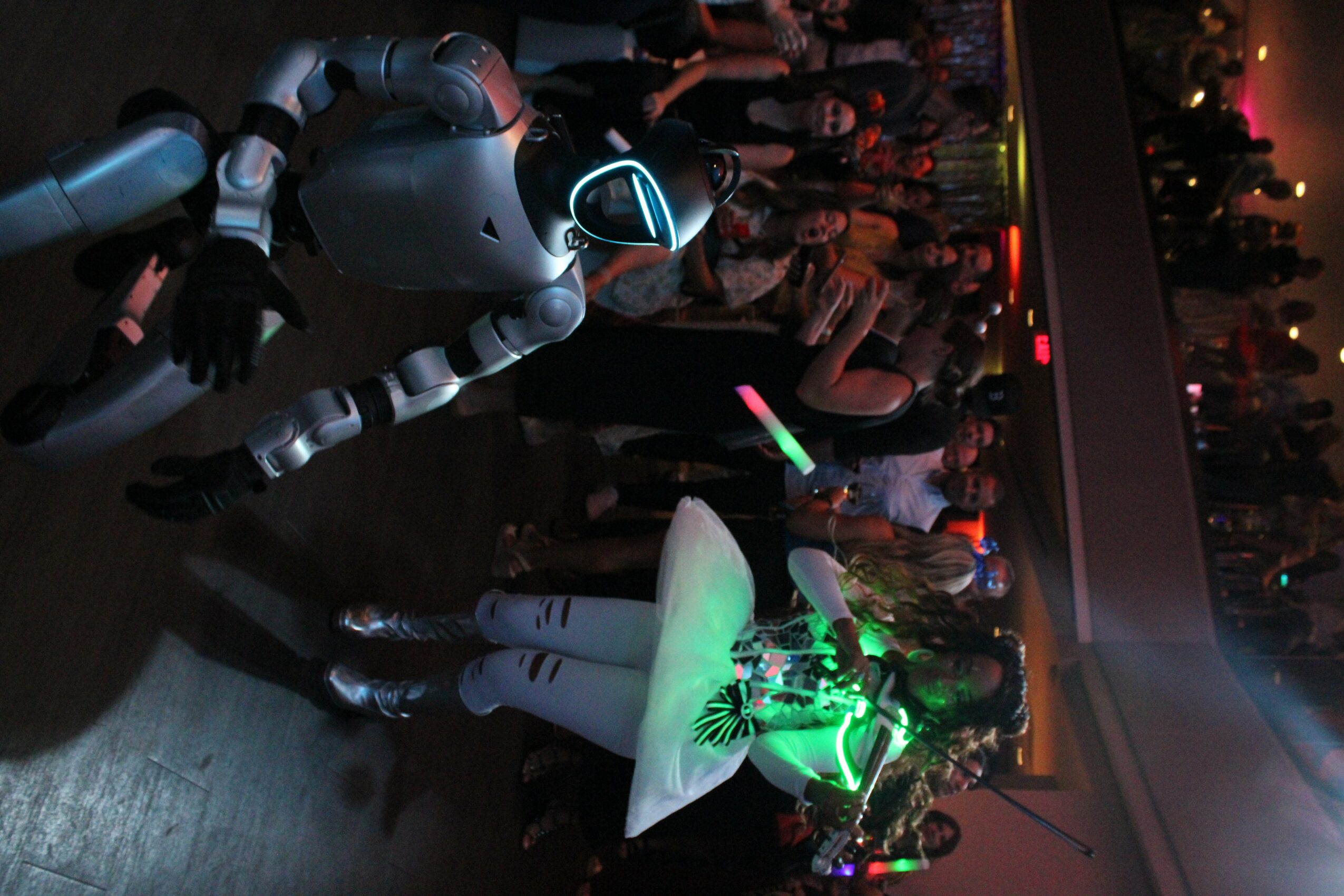 Robot with glowing eyes and a person holding a glowing umbrella at a party with people dancing and colorful lights.