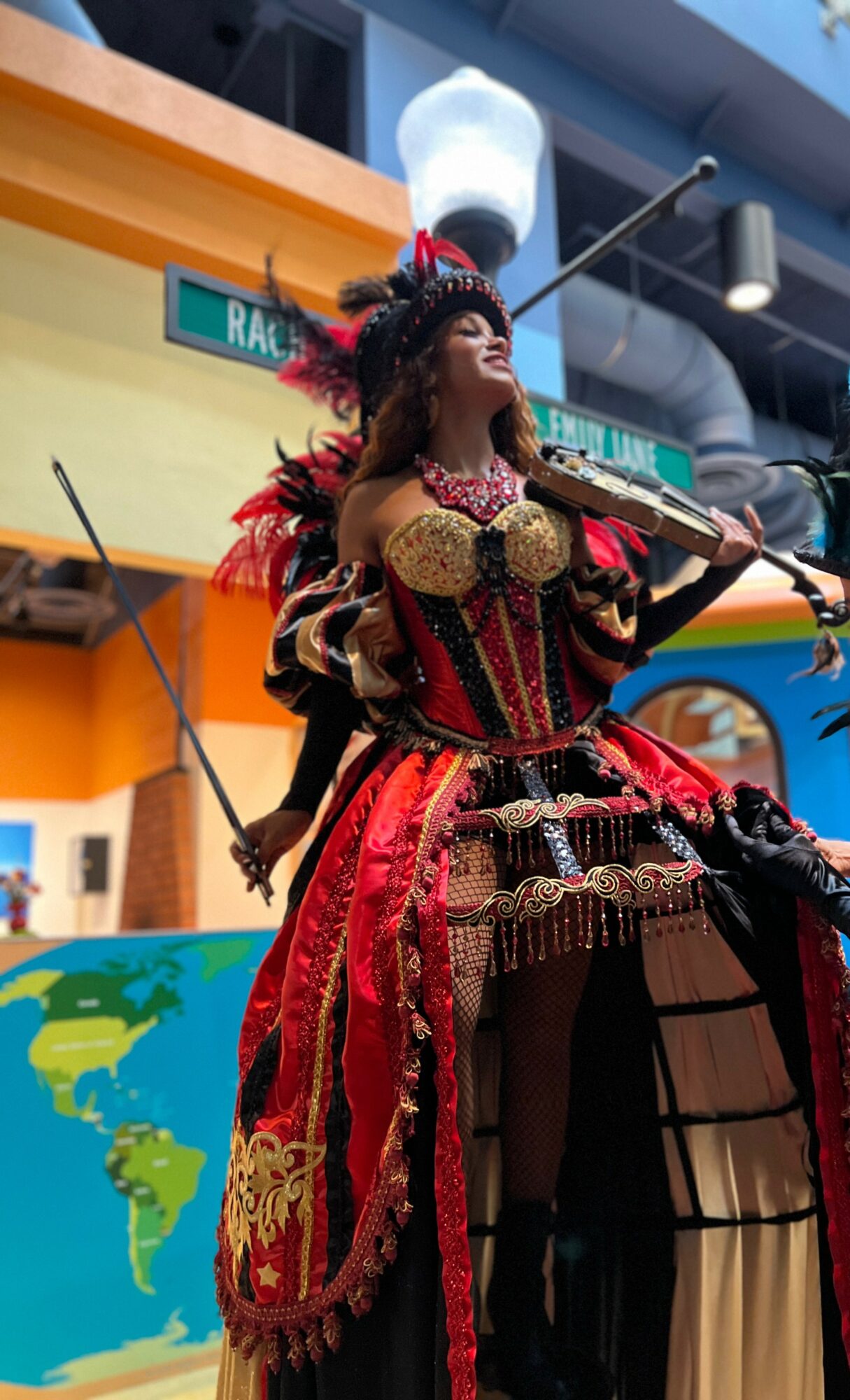 Person in elaborate red and gold costume with feathers, holding a staff, standing outdoors in front of colorful buildings.
