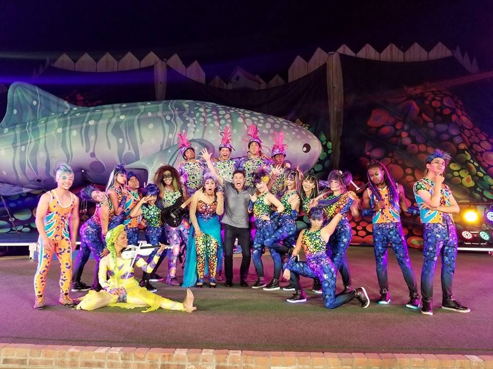 Group of performers in colorful costumes on stage with a large fish prop and vibrant background, posing for a photo.