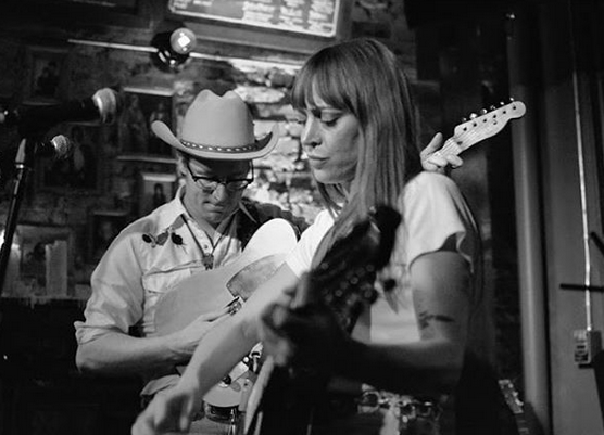 Two musicians, one with a guitar and the other with a microphone, perform on stage in a dimly lit setting.