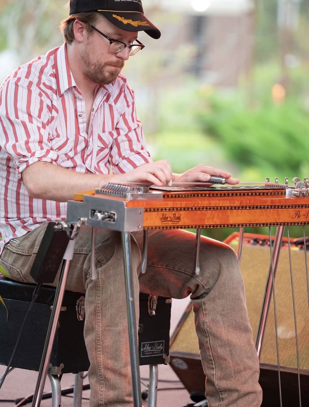 Man playing a steel guitar outdoors, wearing glasses, a striped shirt, and a cap, with a blurred green background.
