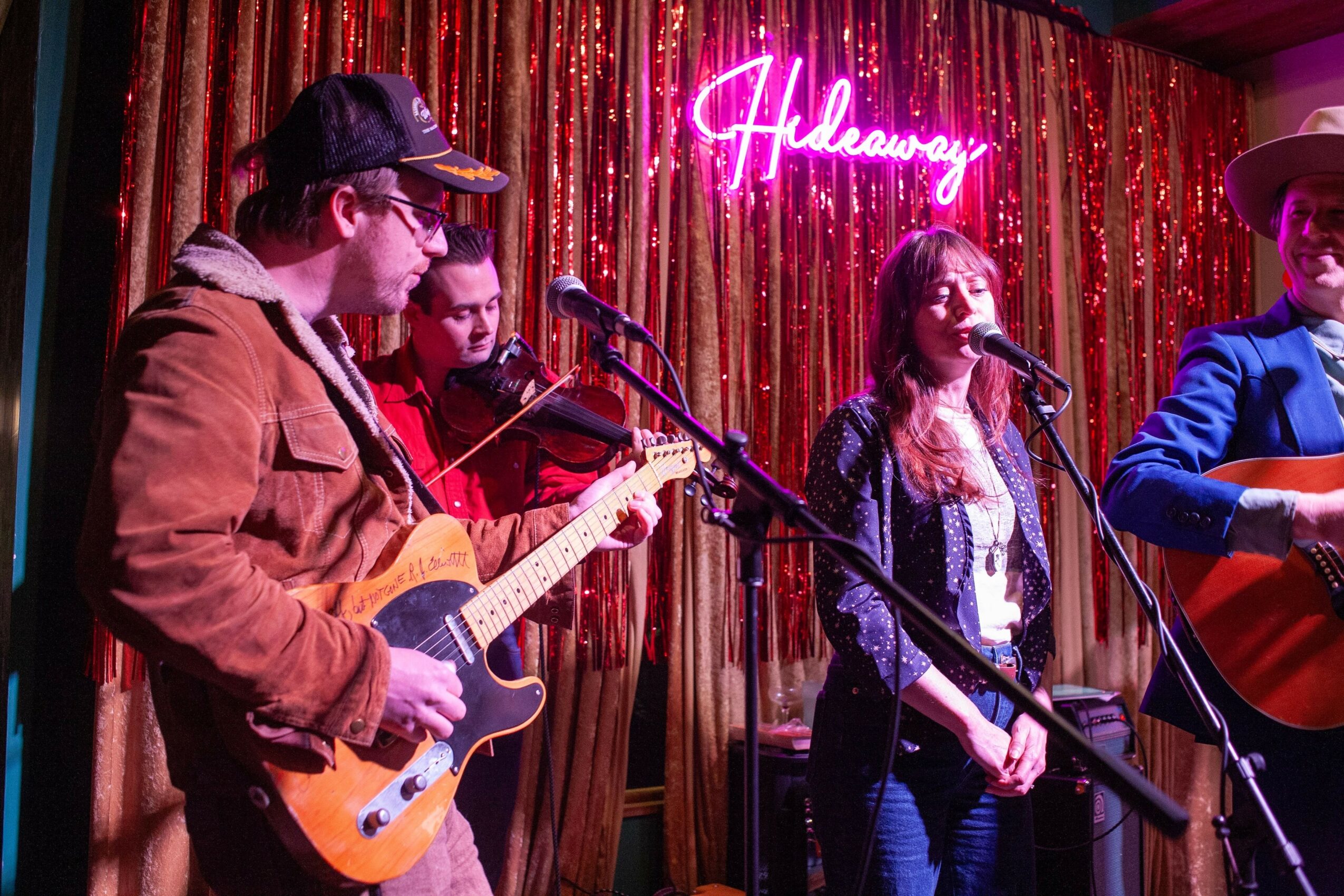 Four musicians perform on stage with a pink neon sign reading 'Halleary' behind them, gold and red curtains in the background.