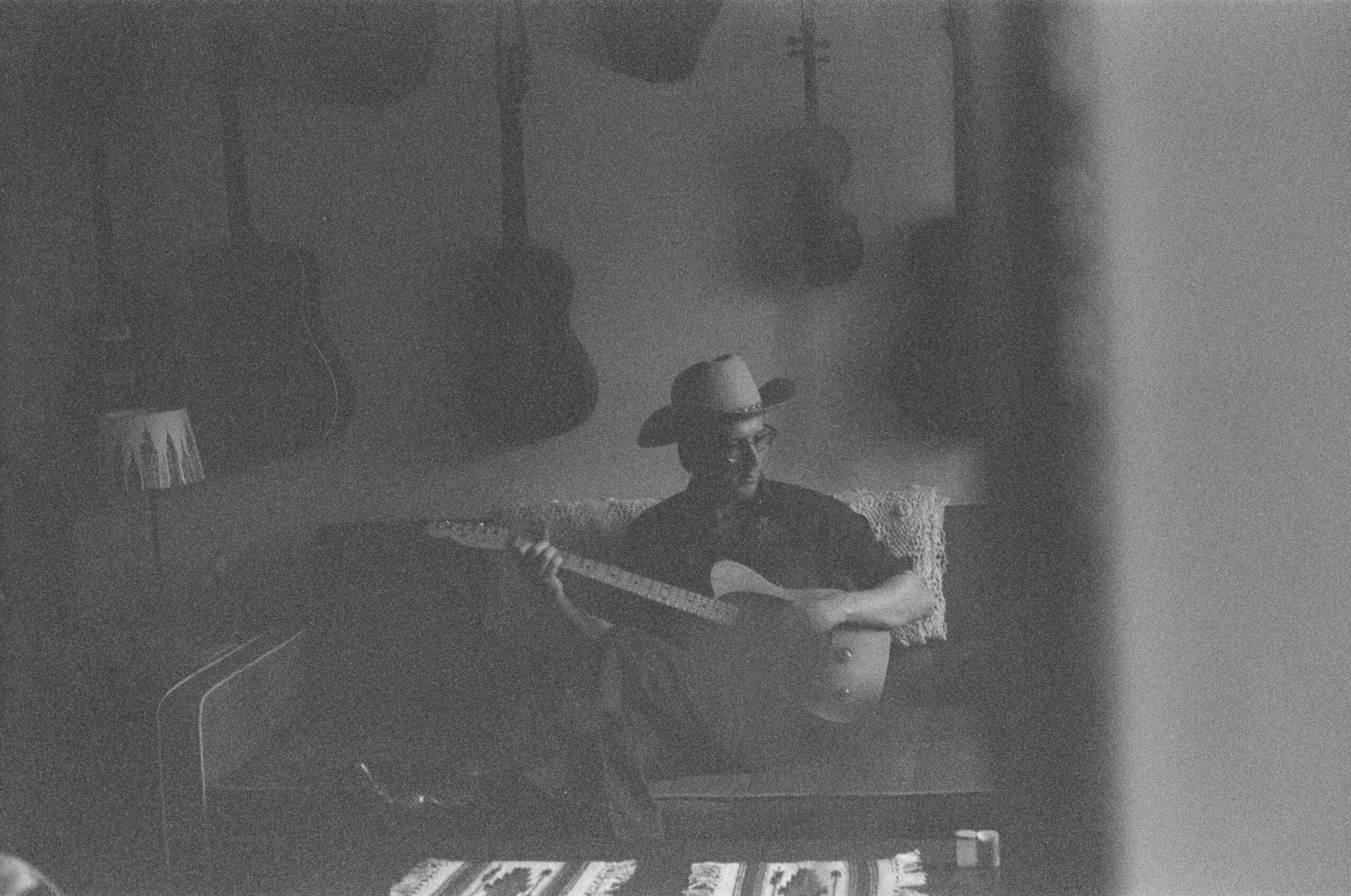 Person wearing a hat playing a guitar in a dimly lit room with guitars hanging on the wall.