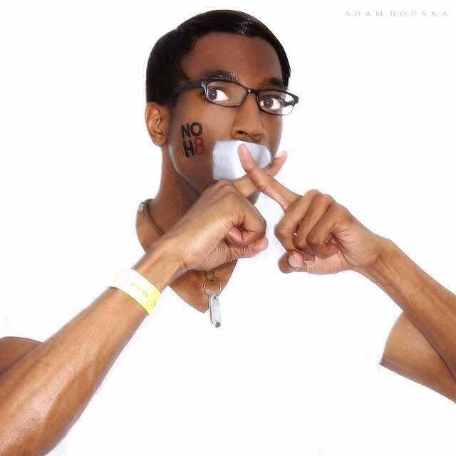 Person with glasses and a wristband, holding a finger to their lips, with a 'NO H8' tattoo on their face, against a white background.
