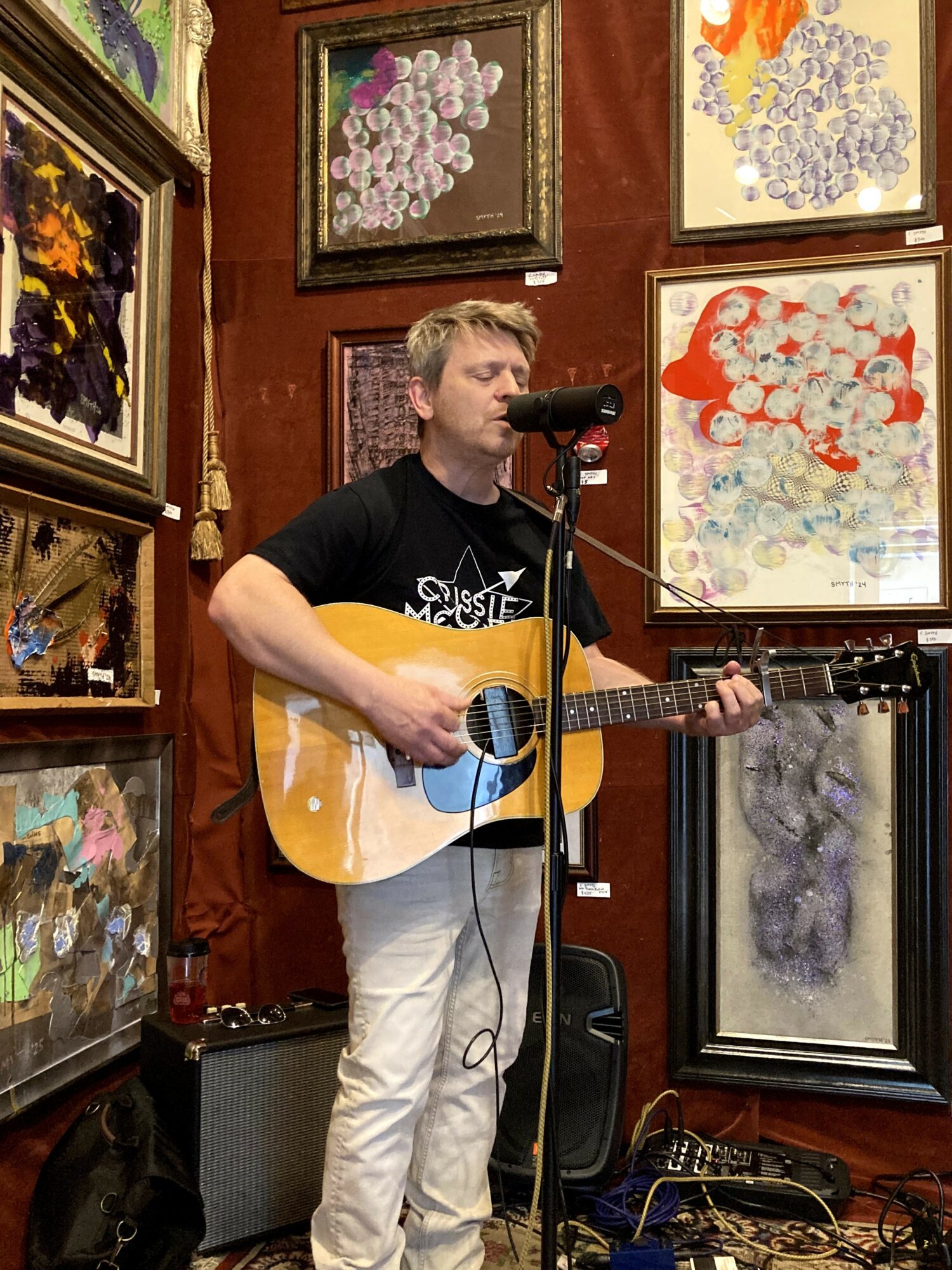Young man with guitar singing into microphone in art gallery with colorful paintings on walls.