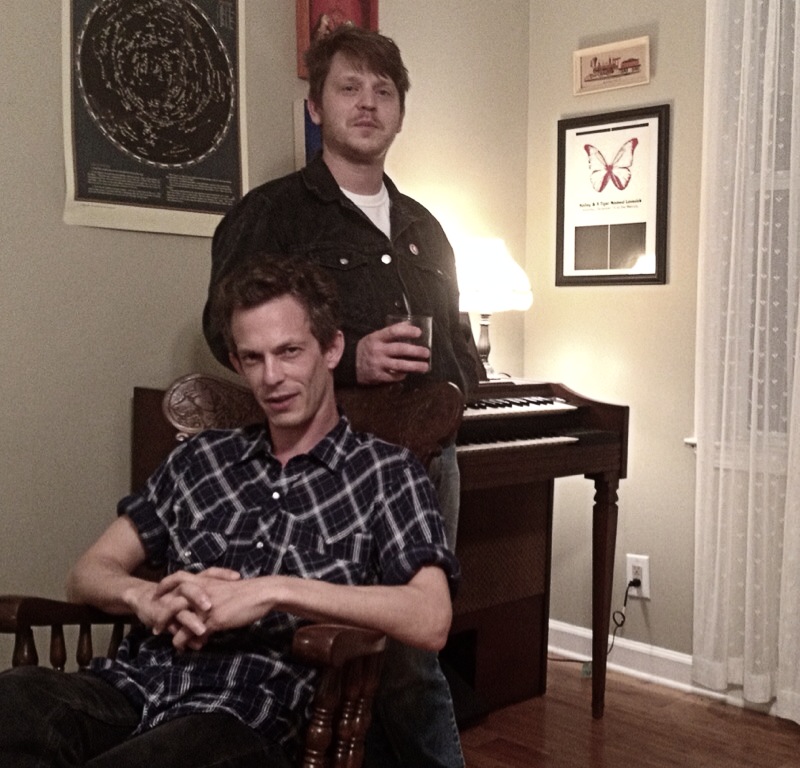 Two men in a room with framed artwork, a piano, and a lamp, one sitting and one standing, holding drinks.