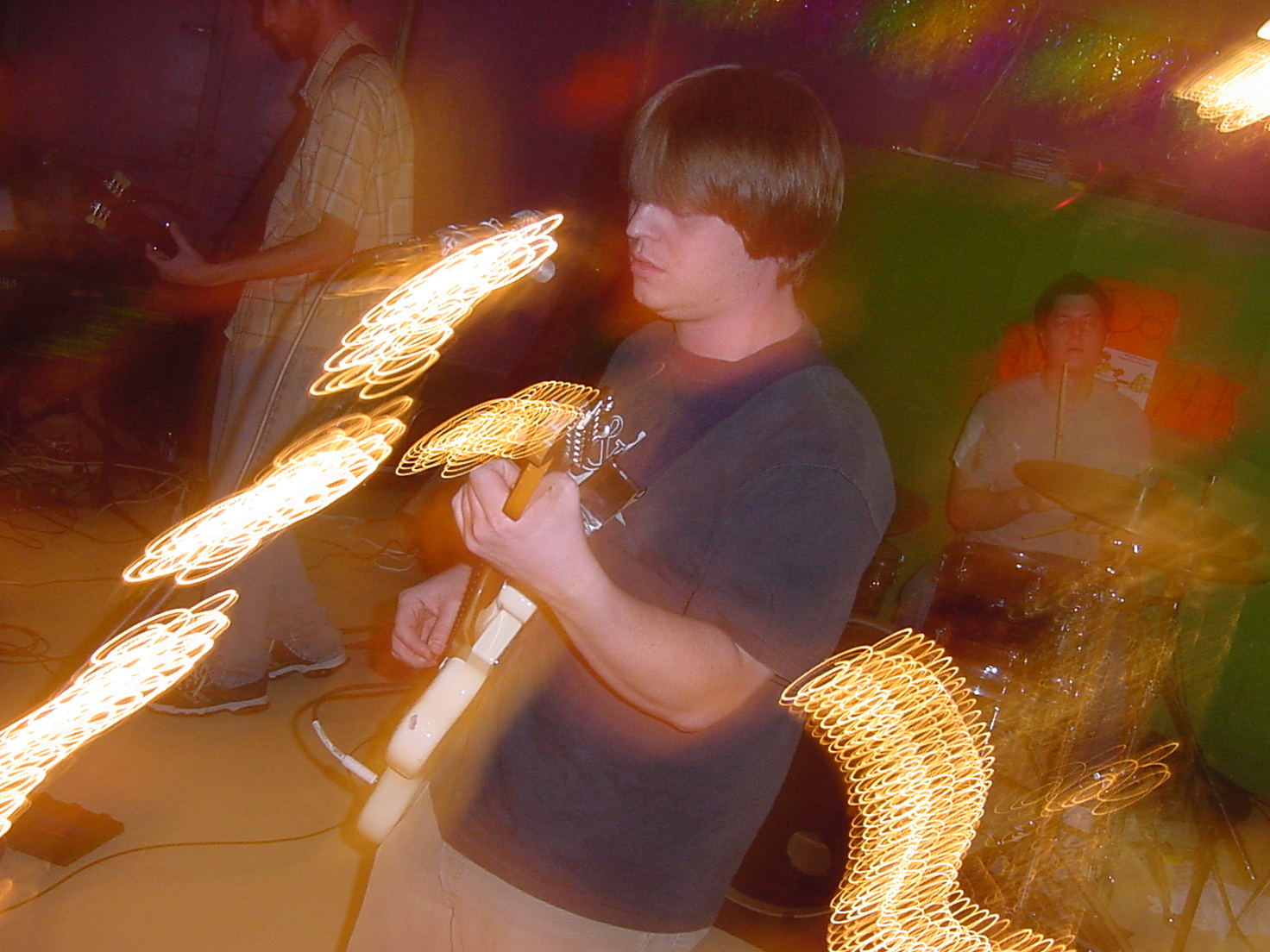 Young person with dark hair holding a guitar, surrounded by colorful lights and blurred background, in a lively setting.