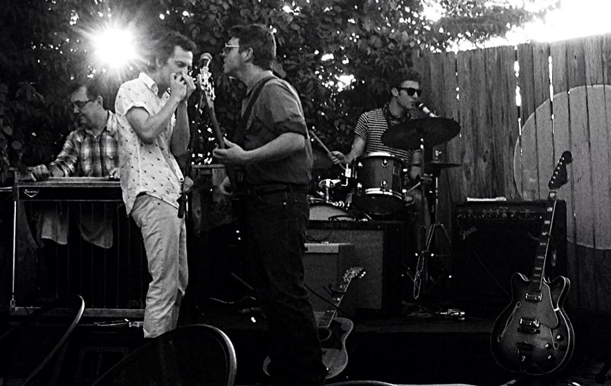 People gathered outdoors, some playing musical instruments, with a wooden fence in the background, sunlight shining through trees.