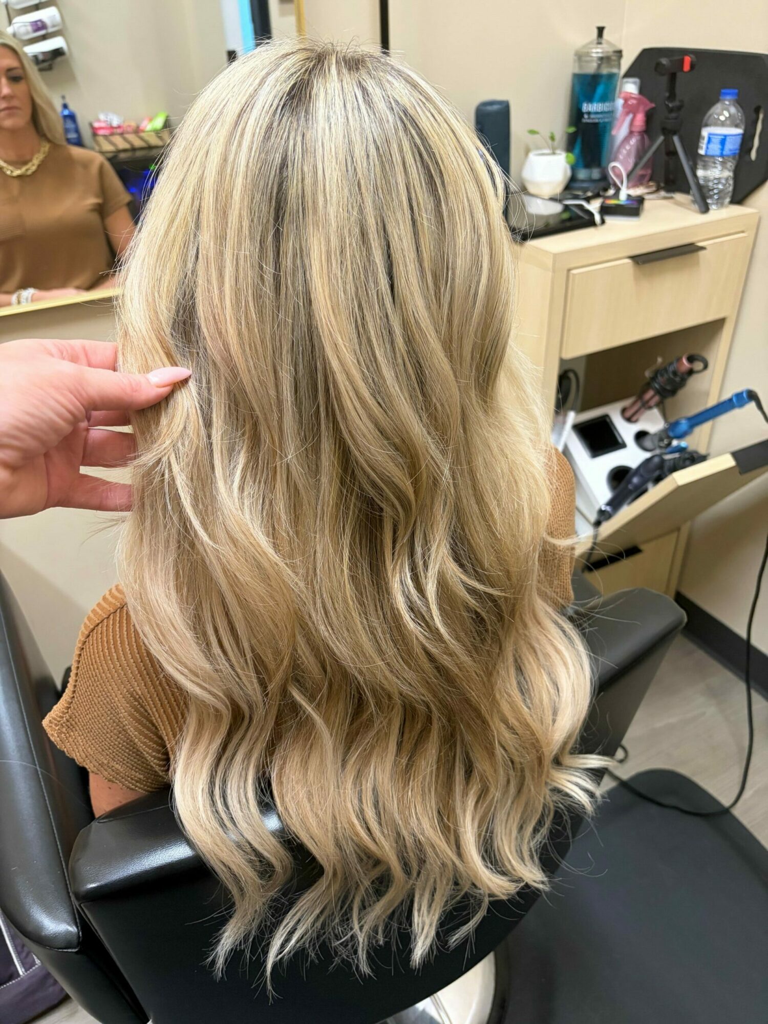 Side view of a woman with long, wavy blonde hair in a salon chair, with salon tools and furniture in background.