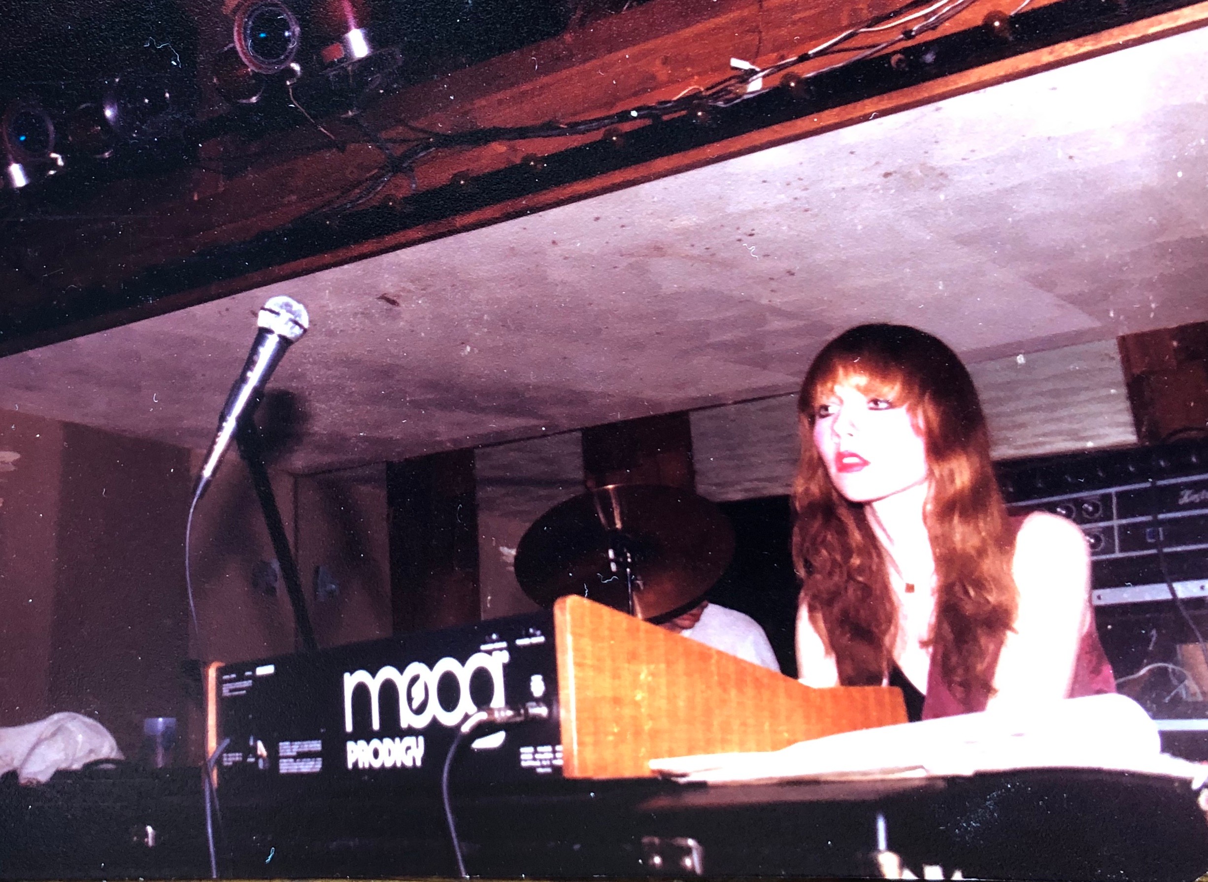 Woman with long hair playing keyboard on stage with microphone nearby, in a dimly lit venue.