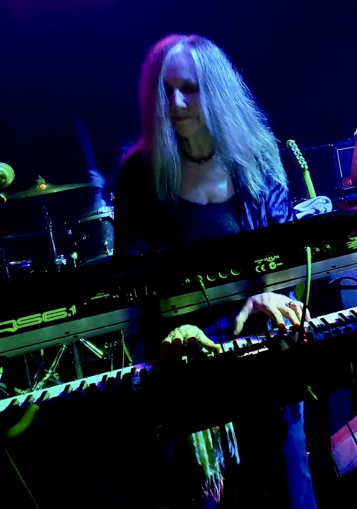 Female musician with long hair playing keyboard on stage with blue and purple lighting.