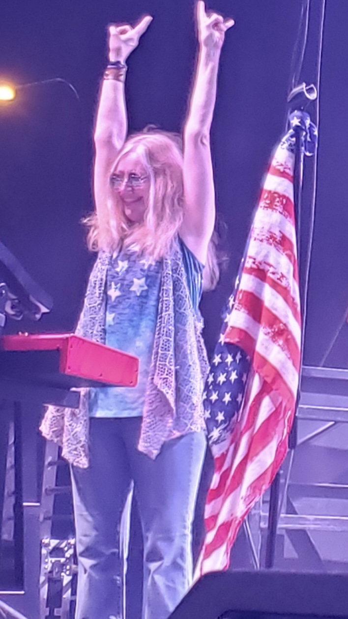 Woman with blonde hair raising both arms, standing next to an American flag, on stage with purple lighting.
