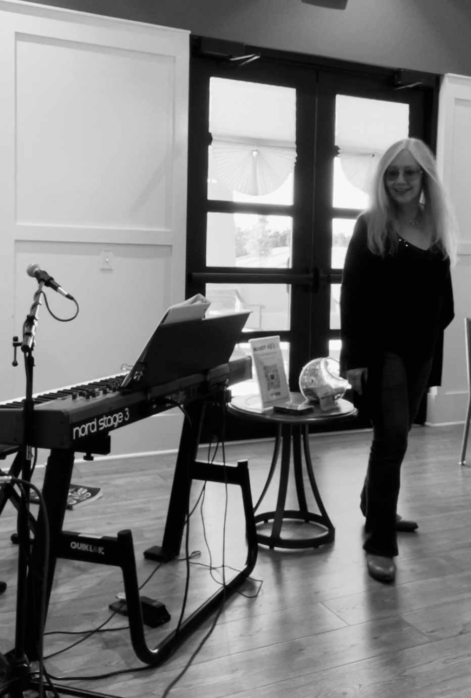 Woman with long hair and glasses standing indoors near a table with a keyboard and a microphone, in front of large windows.
