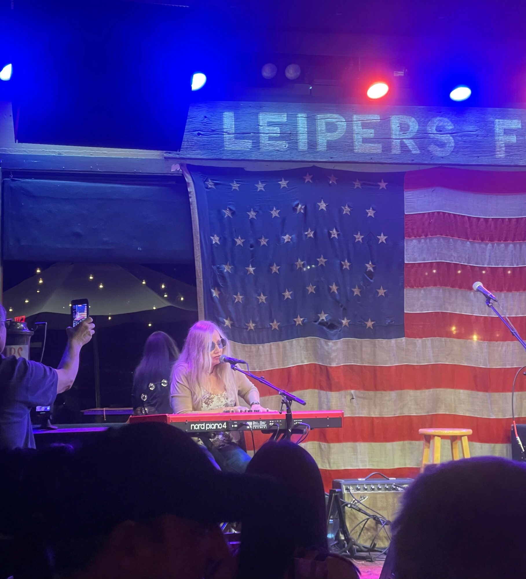 A woman plays a keyboard on stage with an American flag backdrop and a sign reading 'LEIPERS F'.