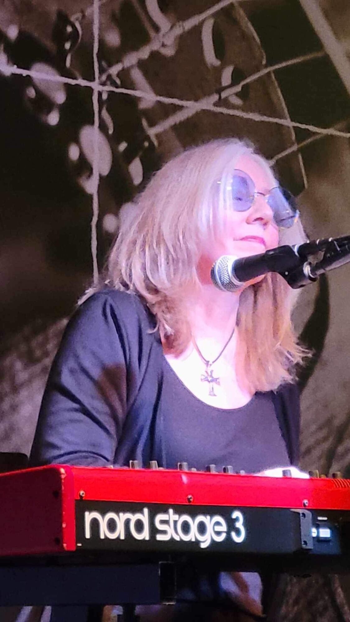 Woman with blonde hair singing into a microphone while playing a red Nord Stage 3 keyboard.