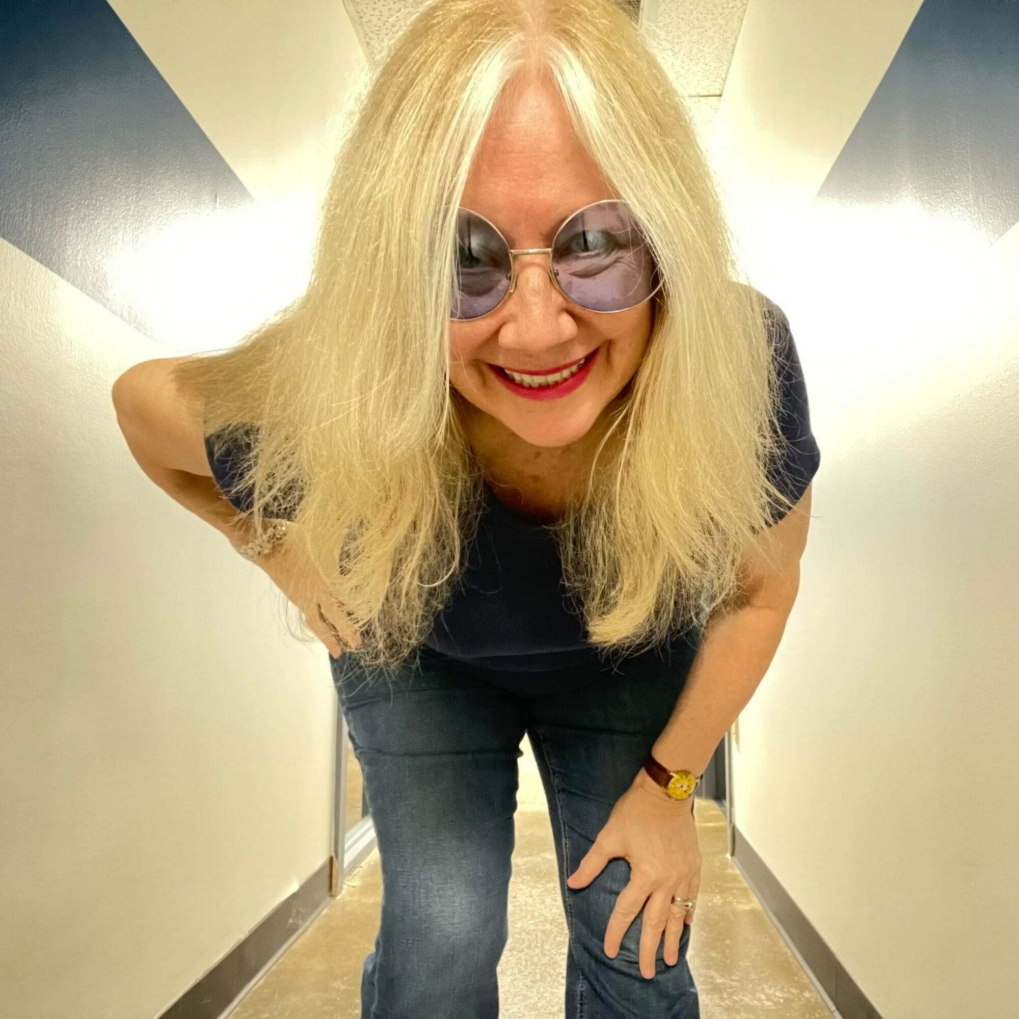 Woman with long blonde hair, sunglasses, smiling, leaning forward in a hallway with colorful walls