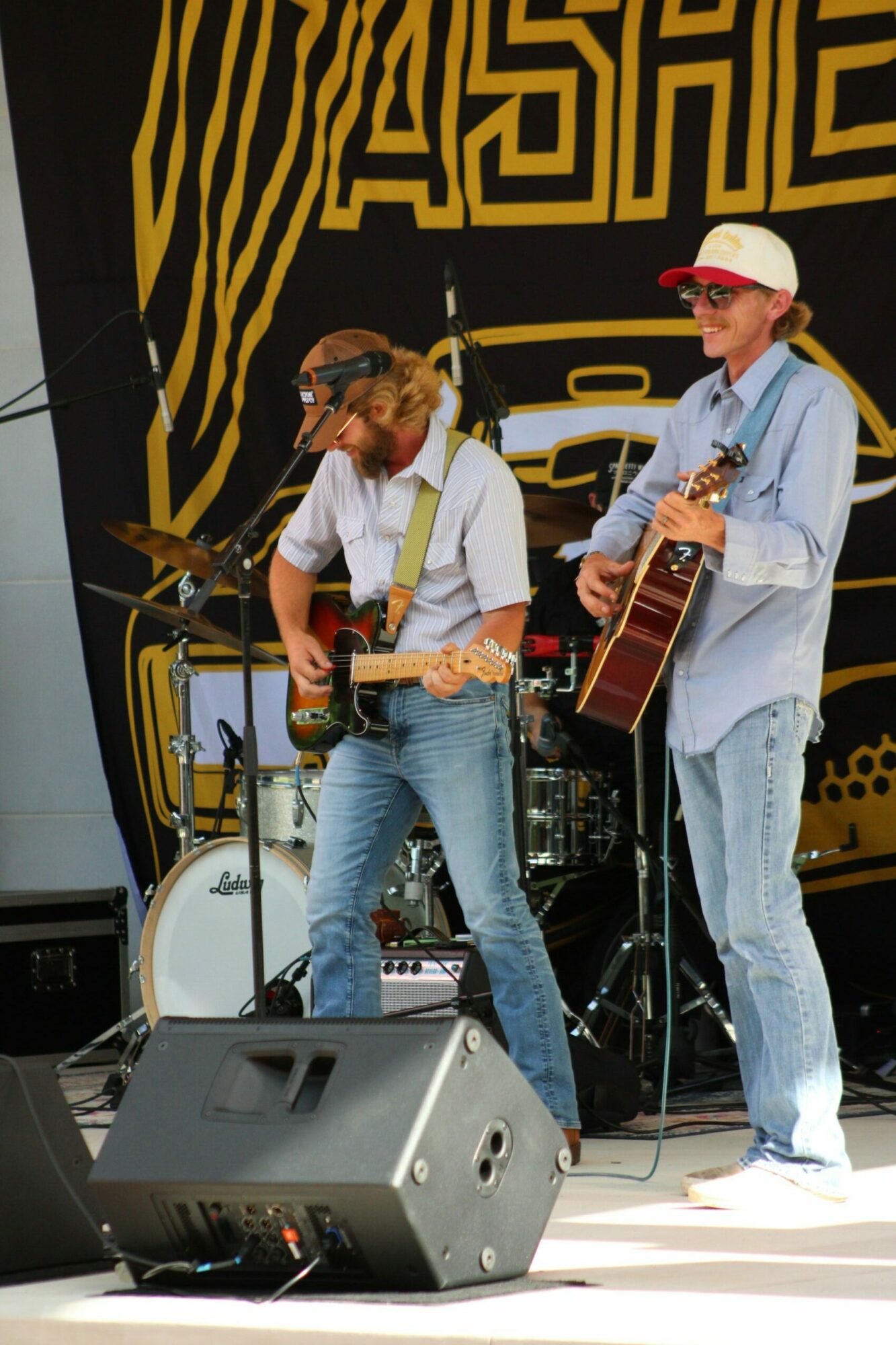 Two musicians perform on stage, one playing guitar and the other singing into a microphone, with a black and yellow backdrop.