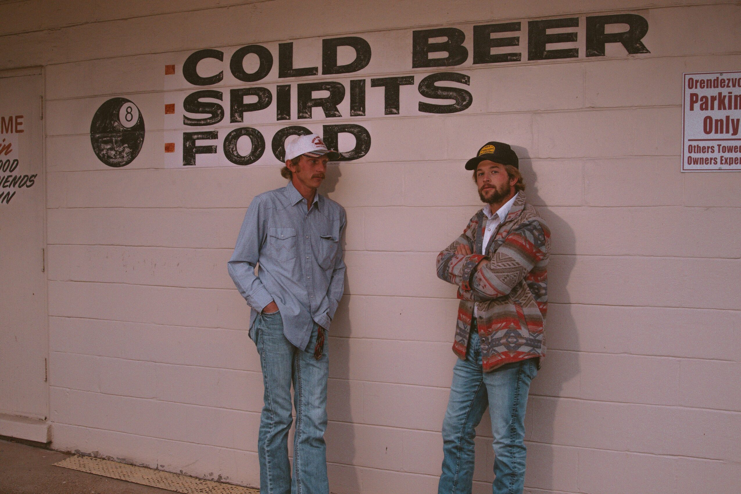 Two men stand against a wall with signs for cold beer, spirits, and food, one wearing a cap and the other with a beard.