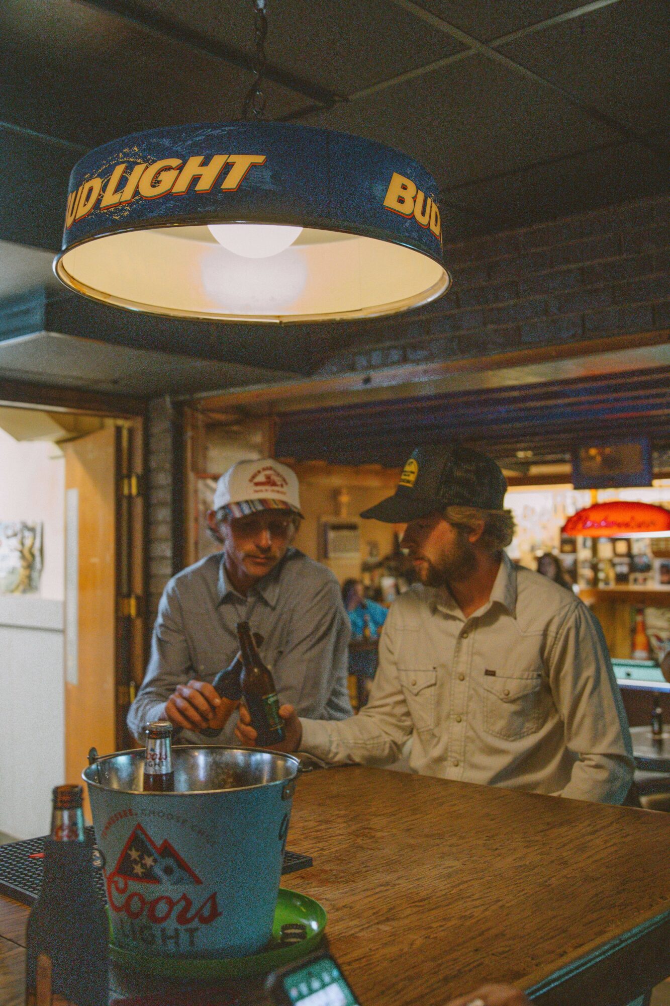 Two men sit at a bar counter, one holding a bottle, with a Coors Light bucket and a menu in front.