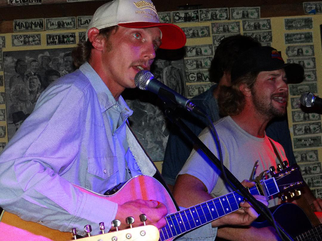 Two men perform with guitars and microphones in a room decorated with dollar bills on the wall.