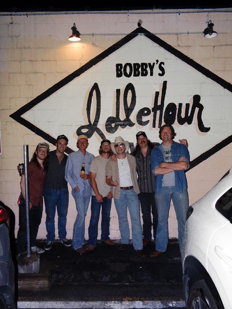 Group of seven people standing in front of a sign that reads 'Bobby's Lighthouse' on a white brick wall.
