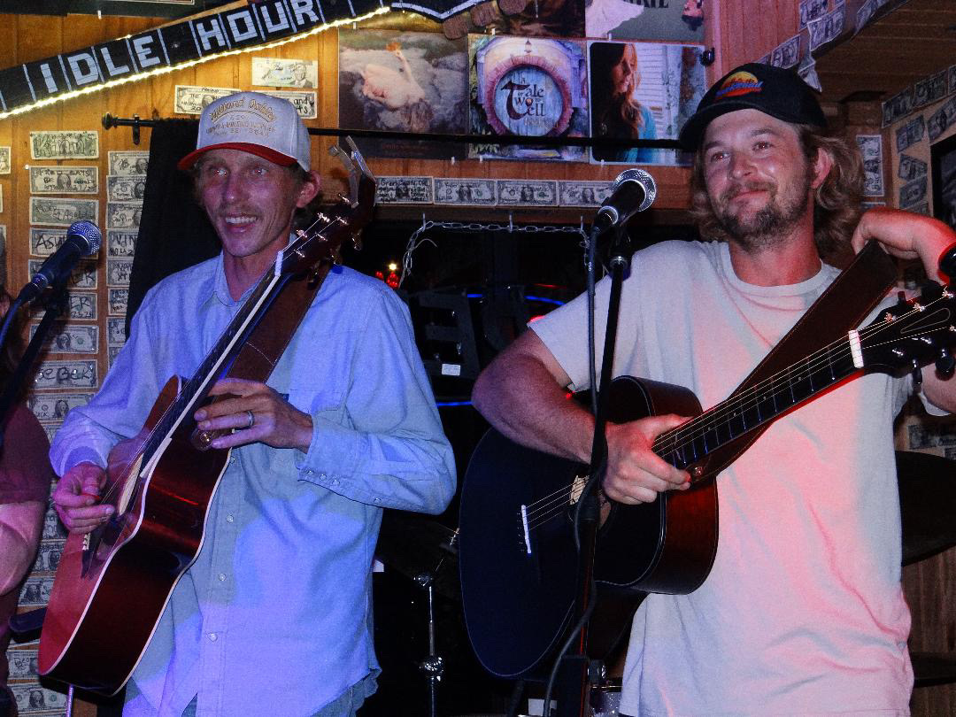 Two men playing guitars and singing into microphones in a bar with brick walls and TV screens behind them.