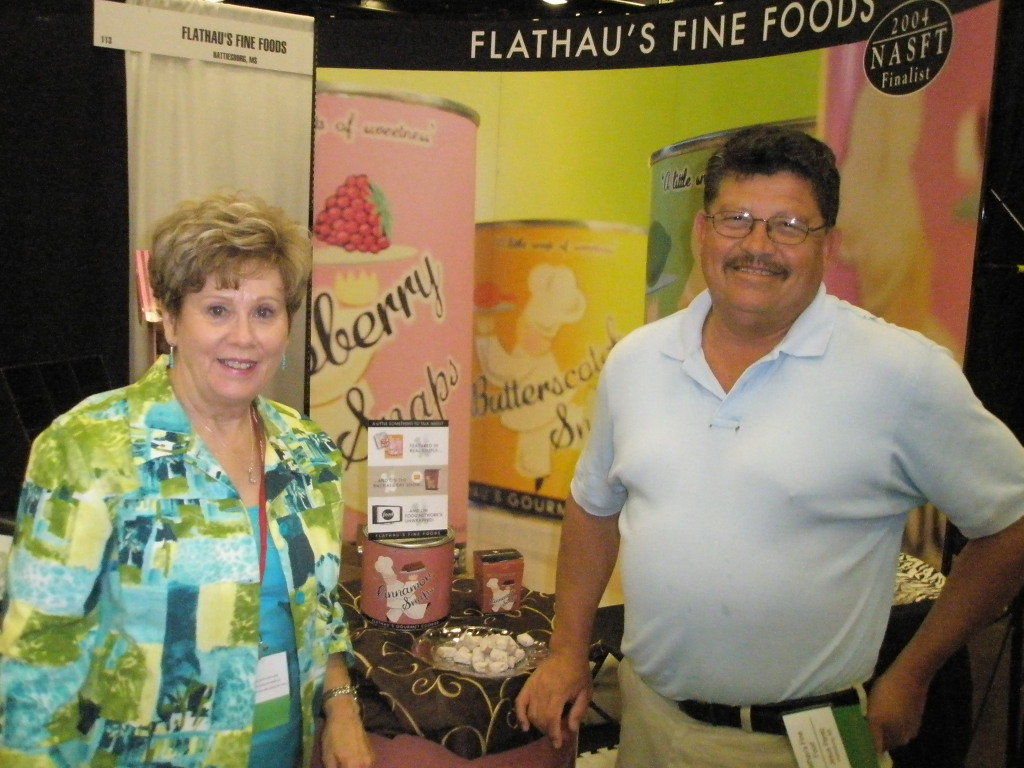 Two people standing in front of a display for Flathau's Fine Foods, with colorful product packaging behind them.