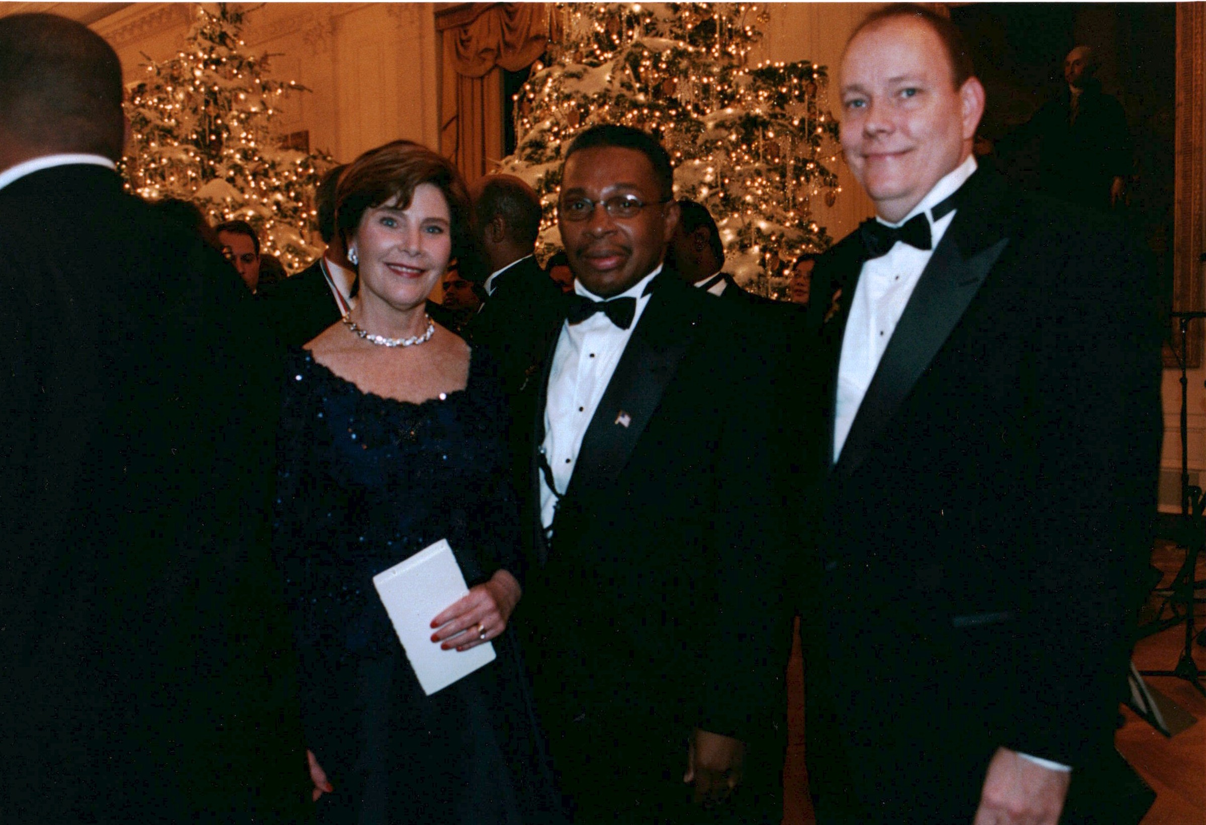 Three people in formal attire at a festive event with decorated Christmas trees in the background.