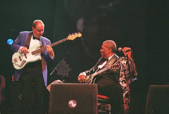 Two musicians perform on stage, one playing bass guitar and the other singing into a microphone, with dark background.