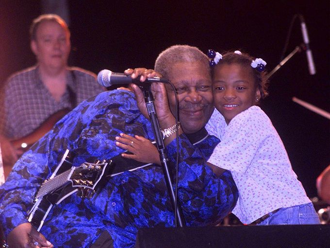 An elderly man with a young girl hugging him, both smiling, on stage with a microphone and a woman in the background.