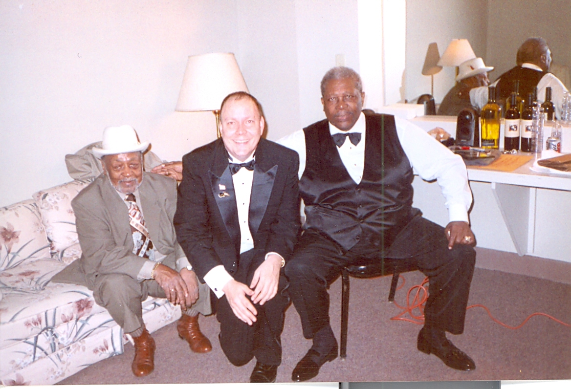 Three men sitting on a sofa and chair in a room with a bar, two lamps, and a mirror, smiling at the camera.
