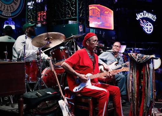 Musician in red outfit playing guitar on stage with drums and other people in background.