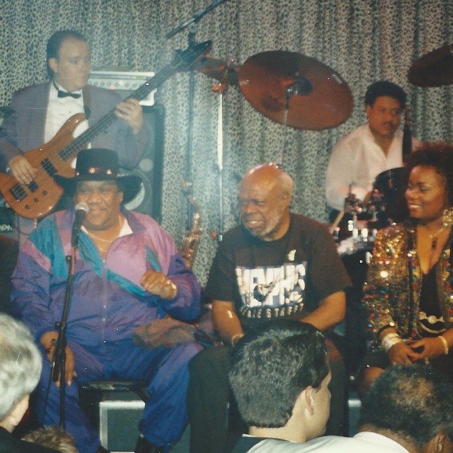 Group of musicians performing on stage with instruments, including a bass guitar, drums, and microphones, in front of an audience.