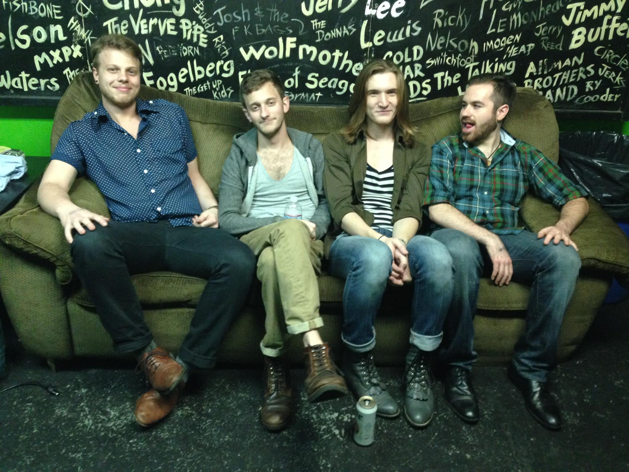 Four people sitting on a green couch in front of a blackboard with names written in white chalk.