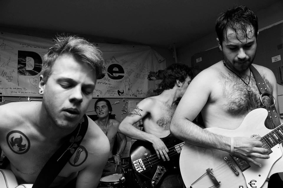 Four shirtless men playing musical instruments in a room with a banner in the background.