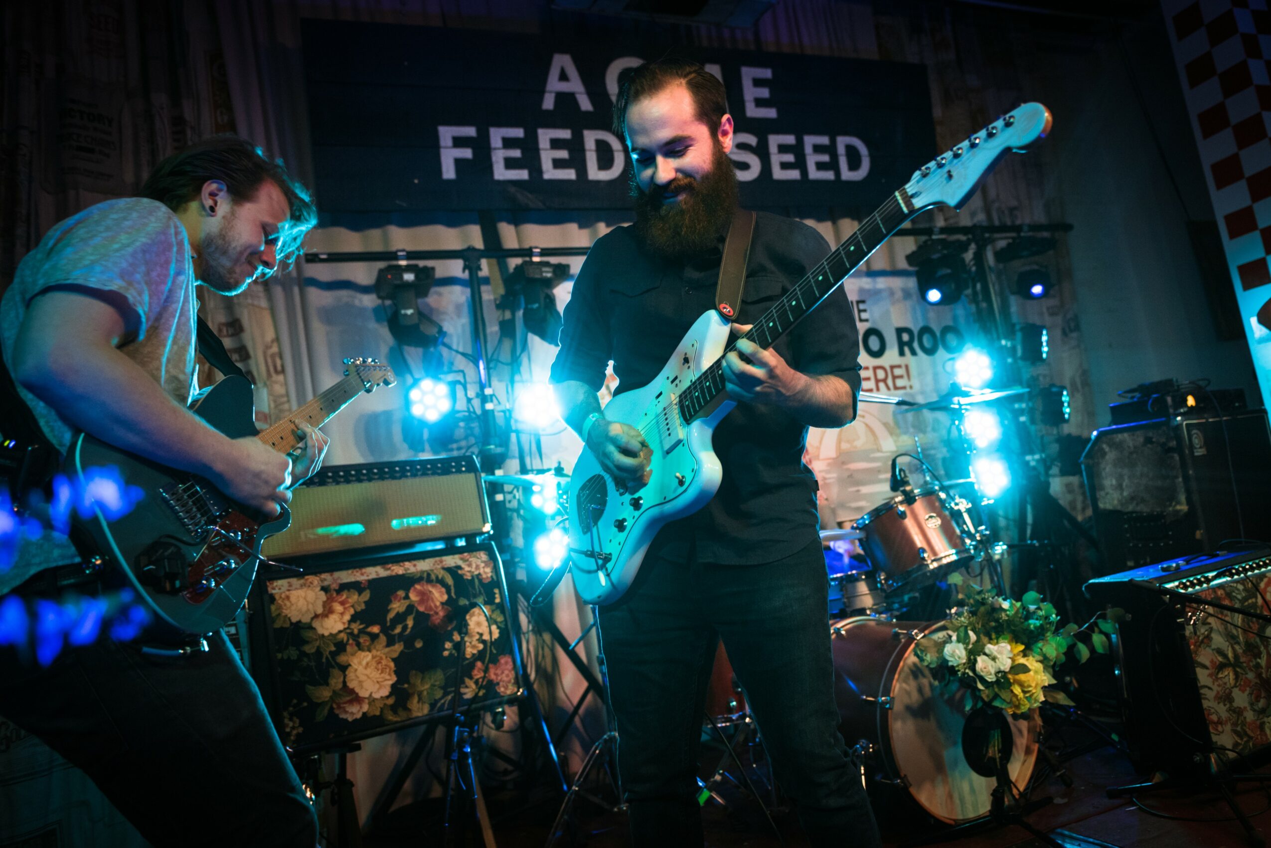 Two musicians playing electric guitars on stage with bright blue lights and a drum set in the background.