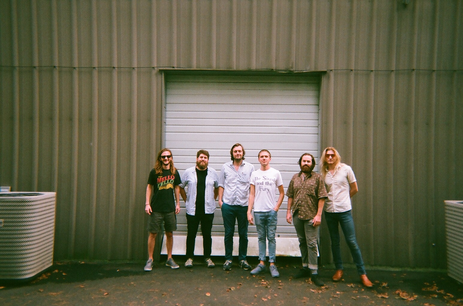 Six people stand in front of a metal building with a closed garage door, outdoors during daytime.